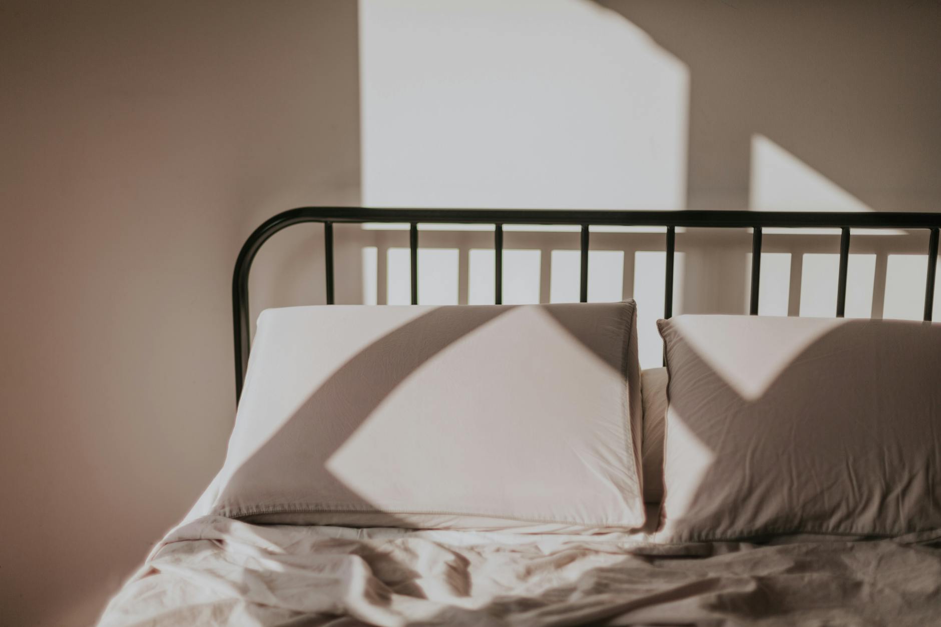 Sunlight casting geometric shadows on a bed with pillows, creating a serene and quiet morning atmosphere.