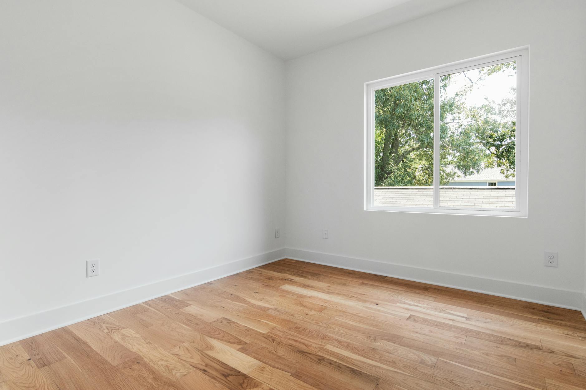Empty room with clean wooden floors and a window overlooking trees, bare walls, natural light