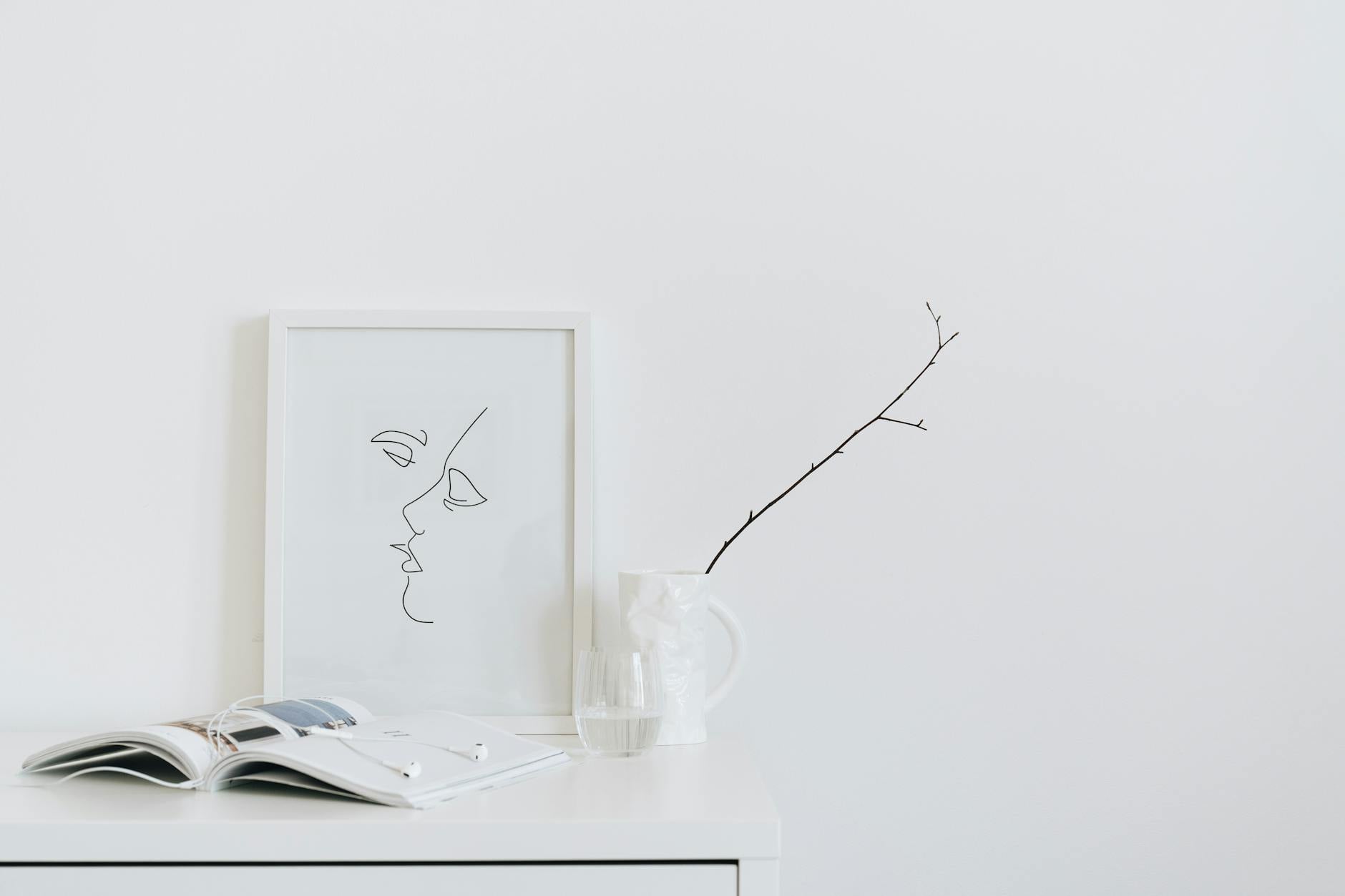 Serene minimalist desk setup with framed art, open book, and glass on a white surface, evoking the calm atmosphere of a thoughtfully designed therapy office