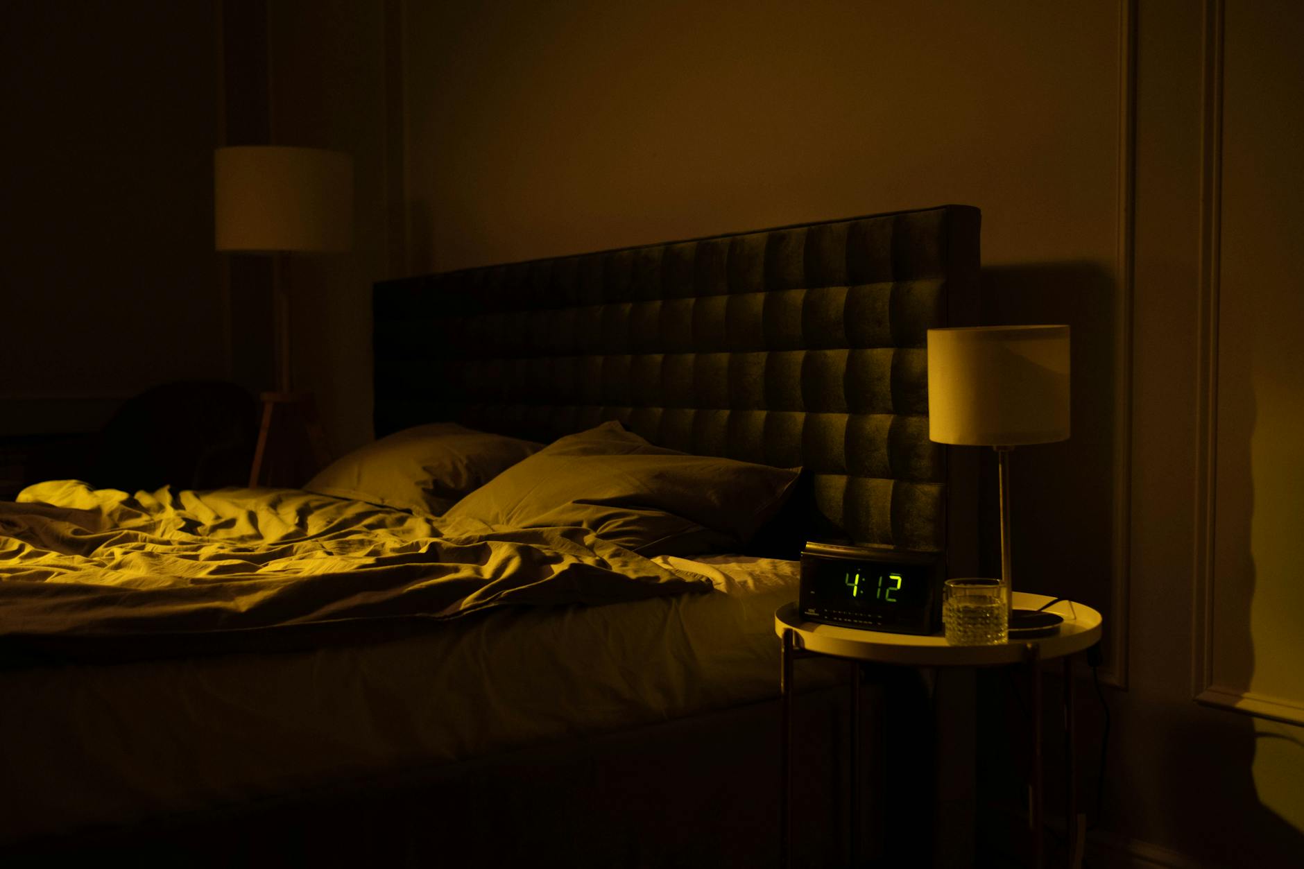 Dimly lit bedroom at night with a digital clock and warm ambient light, unmade bed in the foreground