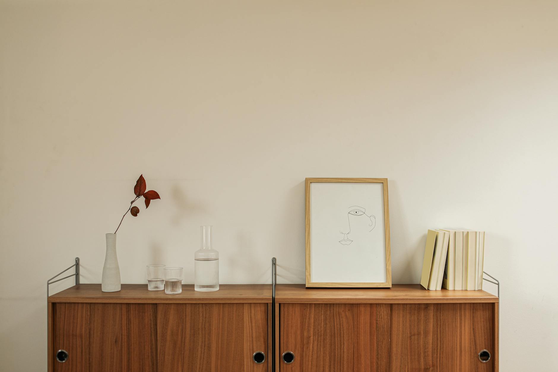 Elegant minimalist interior featuring a wooden cabinet with books, ceramics, and a simple framed art piece — warm neutral tones