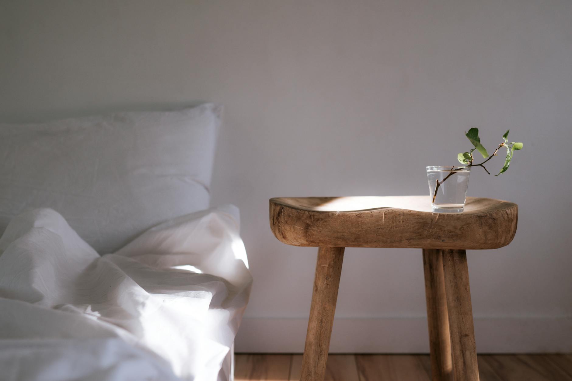 Sunlit minimalist bedroom with white linens and a wooden side table with a plant