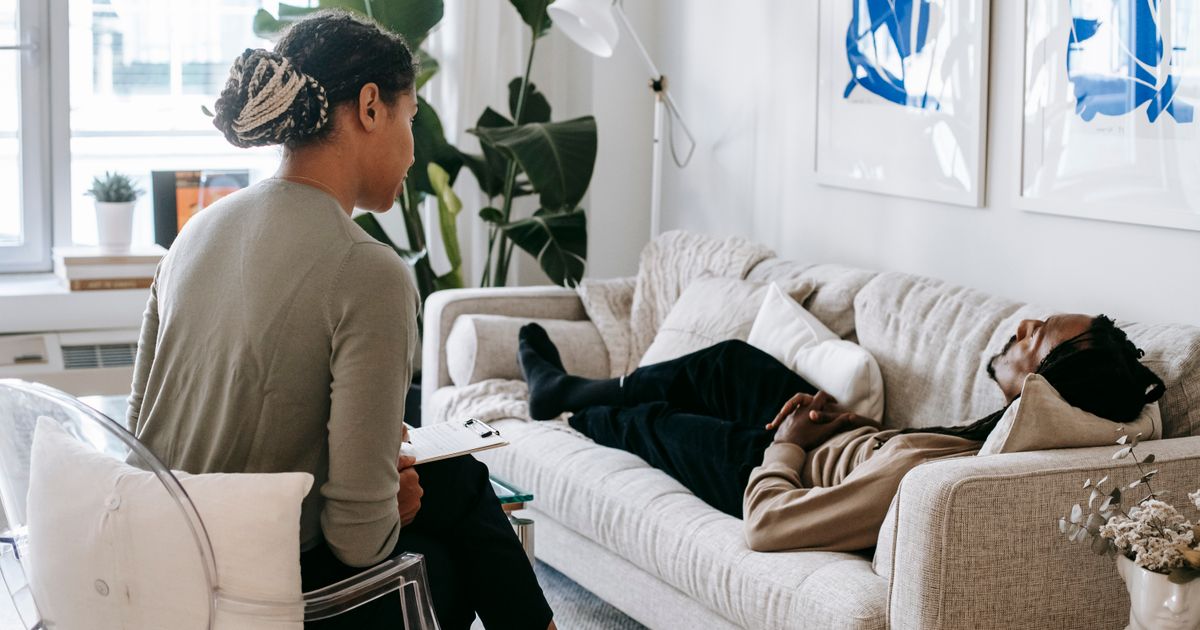 Ethnic female therapist with clipboard sitting near male client lying on couch with clasped hands during psychotherapy session in cozy office