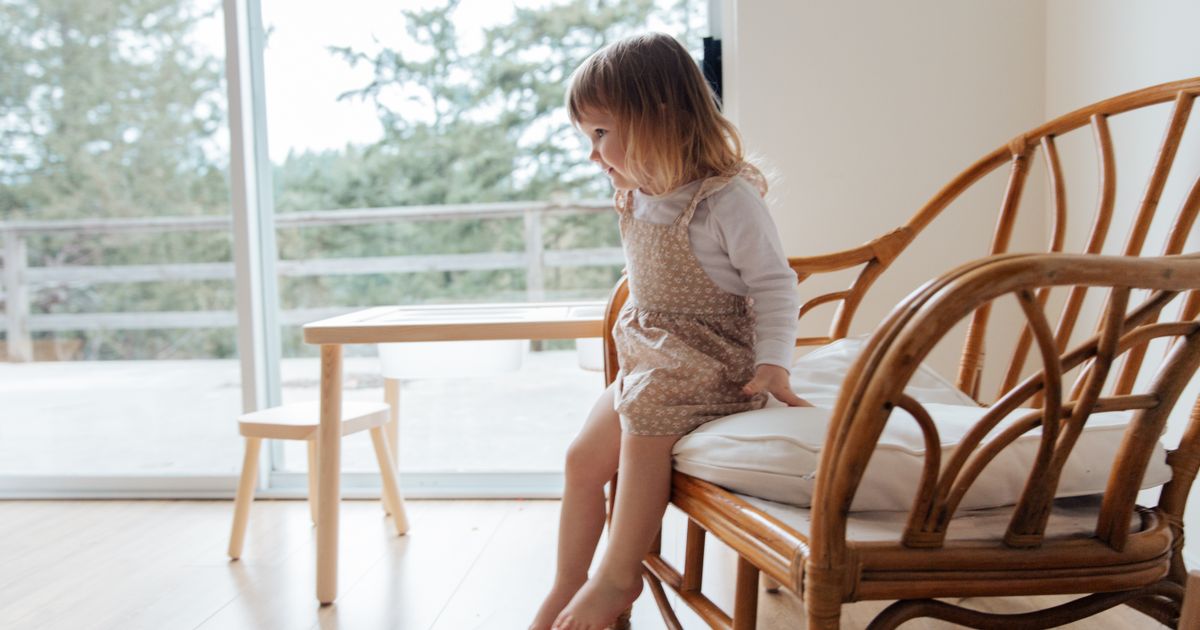 Side view of cute little girl in casual dress sitting on edge of comfortable armchair near table in light living room at home